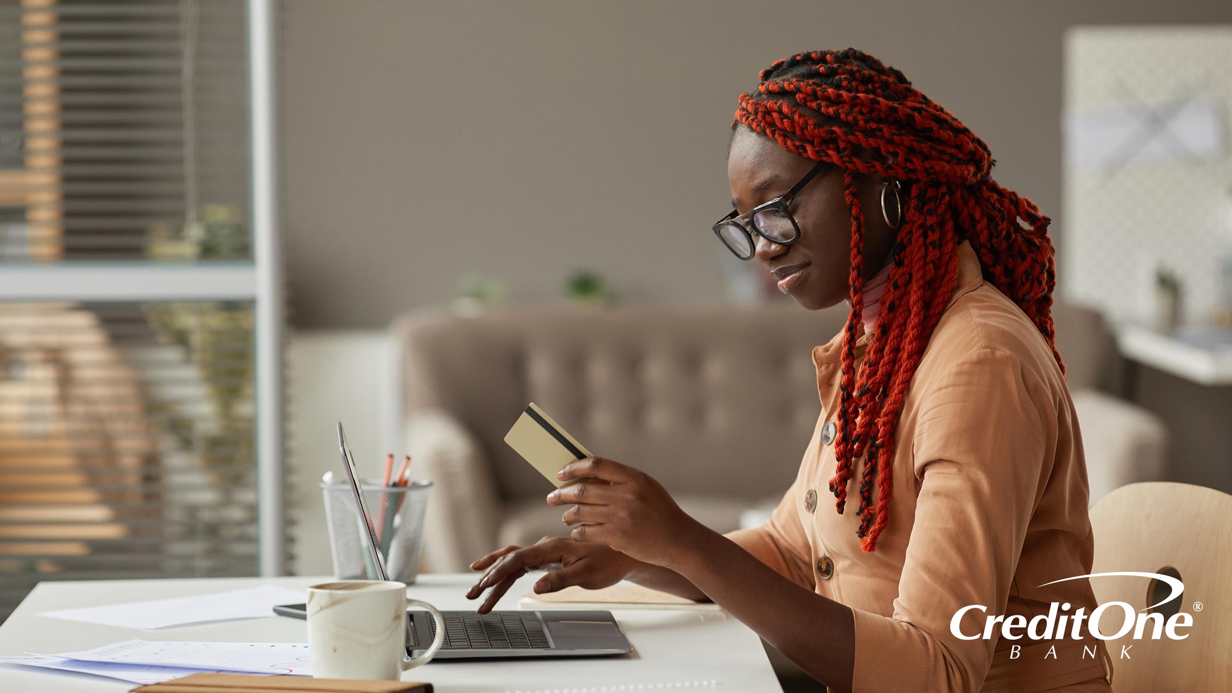 A woman looks at her credit card while using her laptop, taking advantage of the grace period on her credit card for making interest-free online purchases.